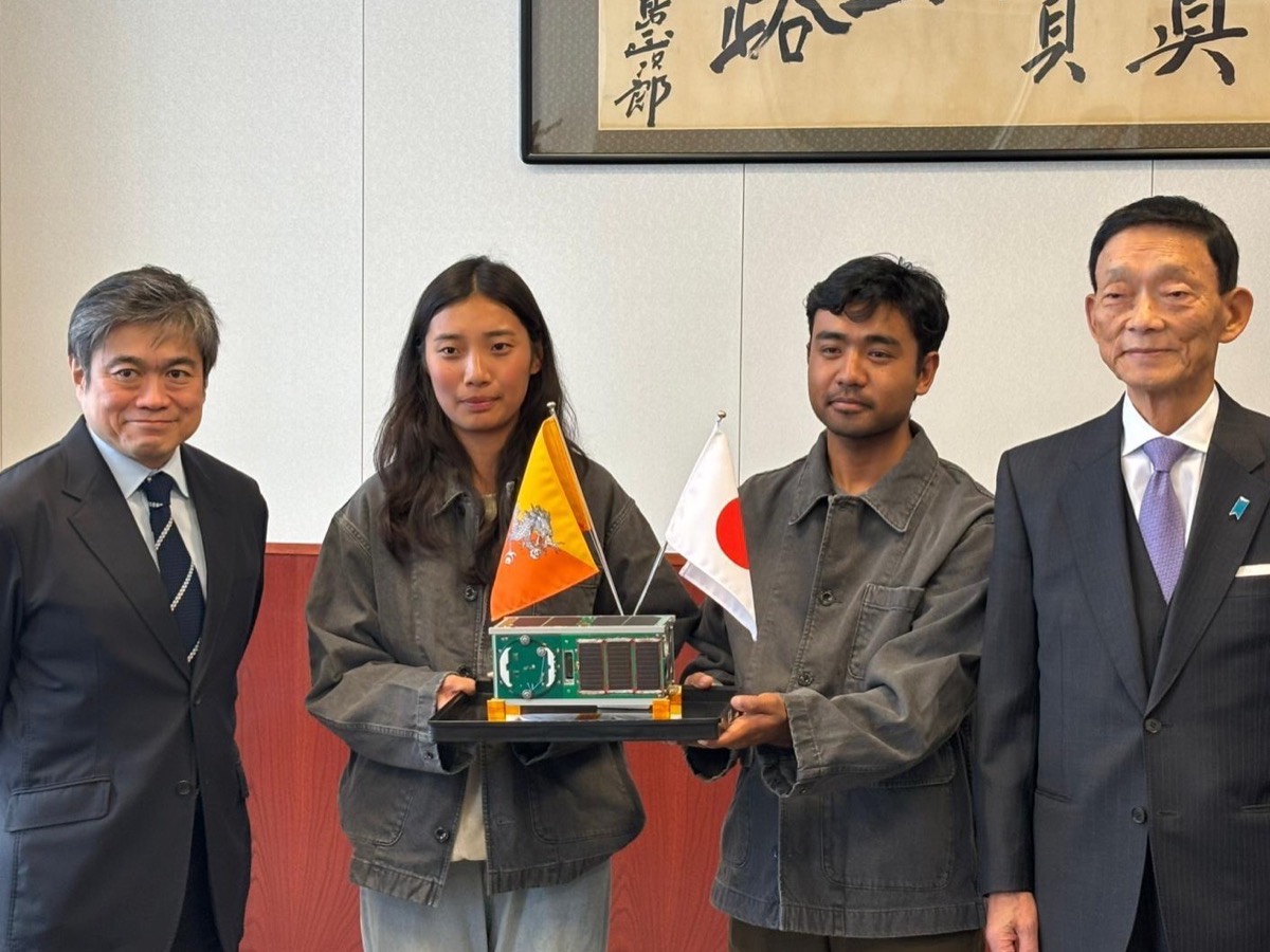 Two students from the College of Science and Technology hold the MOMIJI satellite between the flags of Bhutan and Japan, with Chiba Tech leadership beside them